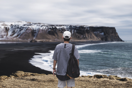 Young man with backpack standing on the cliff at Dyrholaey, Black Sand Beach in Icelandの写真素材