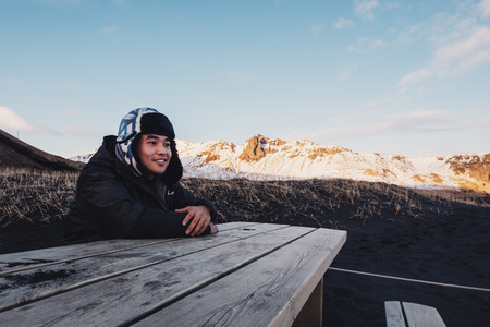 Asian man smiling, sitting on wooden bench in winter at Black Sand Beach in Iceland. traveling Icelandの写真素材