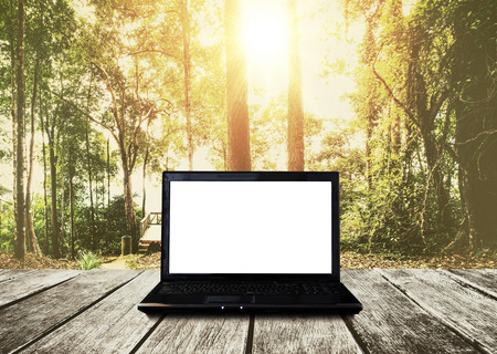 Computer laptop on wooden table, forest in sunrise background. Clipping path empty white screenの写真素材