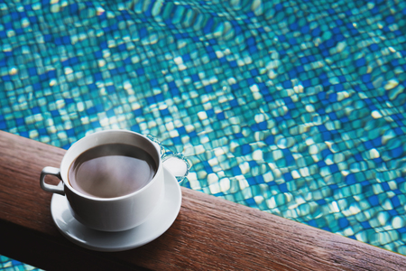 Cup of hot coffee drinks on wood table, with water in swimming pool backgroundの写真素材