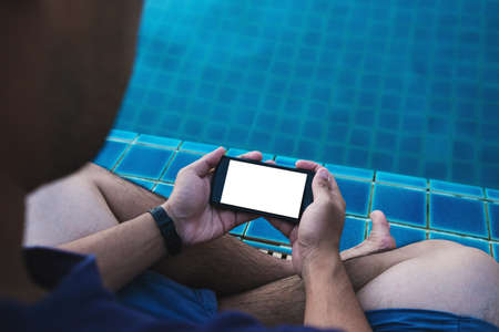 Close-up a man holding mobile smart phone with blank copy space on screen, with legs dipping in pool in summer for vacation. Clipping path on mobile phone screenの写真素材