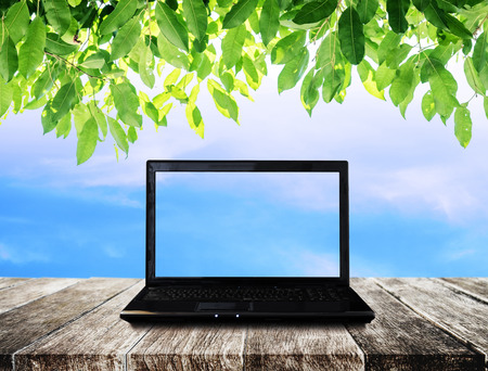 Laptop computer on wooden table with blue sky and green leaves backgroundの写真素材