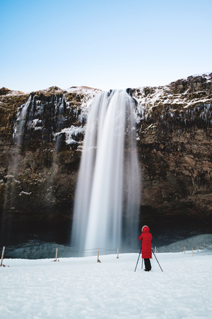 Waterfall in winter, photographer in red jacket standing alone on the snow at Seljalandsfoss waterfall in Icelandの写真素材