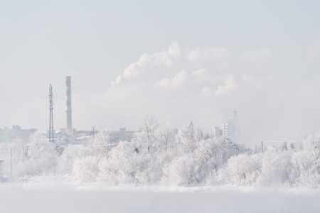 Snow covered on trees with industrial factory and smoke on the sky, white forest landscapeの写真素材