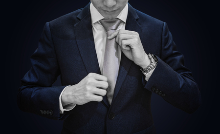 Businessman in dark blue suit tying grey necktie on blue backgroundの写真素材