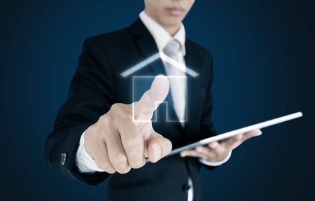 Businessman holding digital tablet and touching house icon on screen, on blue backgroundの写真素材