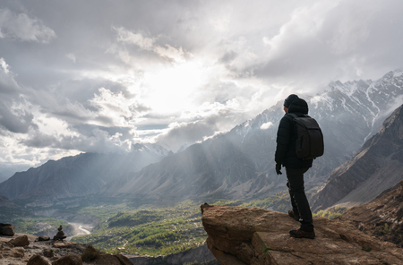 a man with backpack standing on mountain peak and bright sunlight through cloudy sky. Travel lifestyle and achievement success conceptの写真素材