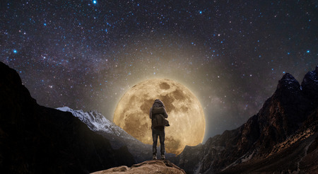 a man with backpack standing on mountain peak at night, and silhouette mountain with bright full moon and sky full of starsの写真素材