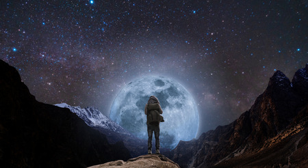 a man with backpack standing on mountain peak at night, and silhouette mountain with bright full moon and sky full of starsの写真素材