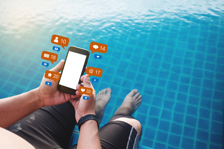 a man using mobile phone at poolside in summer with social media and online message notification icons, blank white screenの写真素材