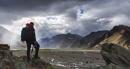 a man with backpack holding camera standing on cliff with mountains view and sunlight through cloud. Travel lifestyle, adventure and success conceptの写真素材