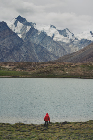 Traveller man in red jacket standing on meadow with lake and snow mountain view. Travel lifestyle, adventure and success conceptの写真素材