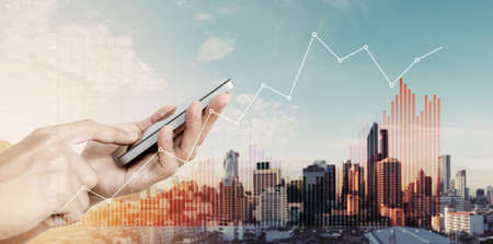 Hand holding mobile smart phone with raising financial graphs, on city backgroundの写真素材