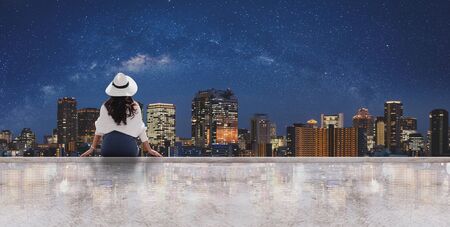 Woman in white hat and clothes sitting on rooftop floor, looking Osaka city at night with sky full of starsの写真素材