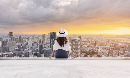 Woman in white hat and clothes sitting on rooftop floor, looking at Osaka city in sunsetの写真素材