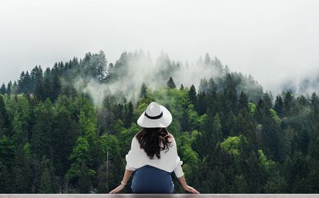 Woman in white hat looking at misty landscape with pine forests in the morningの写真素材