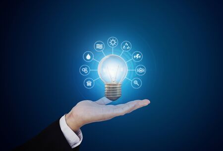 Hand holding bright light bulb with energy resources icon, on blue background. Energy conceptの写真素材