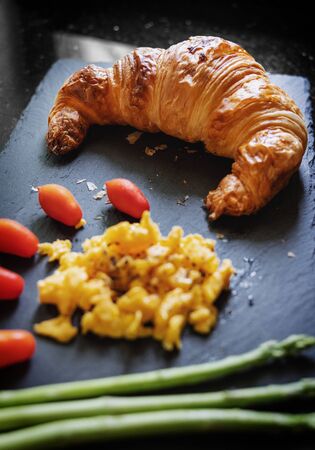 Fresh Croissant with scrambled eggs, cherry tomatoes and Asparagus on black stone plateの写真素材