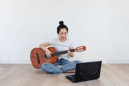 Young Asian woman play guitar, with computer laptop in living room. Home hobbies and recreation, Online training and learning classの写真素材