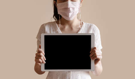 Young woman wearing medical face mask, and showing digital tablet with blank black screenの写真素材