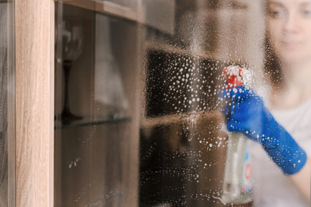a girl in blue gloves cleans the glass with a blue rag, sprinkles with a cleaning agentの写真素材