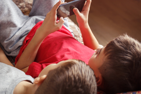 the children are smiling, looking at the display of the mobile phone. Two brothers lying playing a gameの写真素材