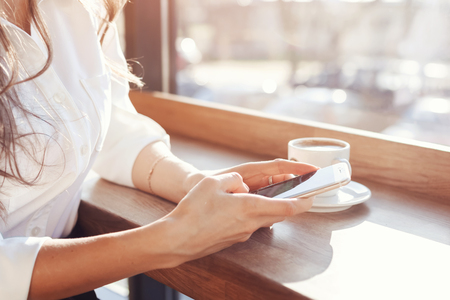 a girl in a white blouse is sitting at a wooden table in a cafe, drinking coffee and holding a phone.の写真素材