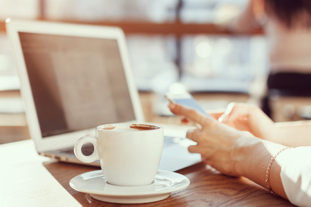 woman working in a cafe for a laptop and drinking coffee, sitting at a table, looking at the phone, in the foreground a cup of coffee. Toned photoの写真素材