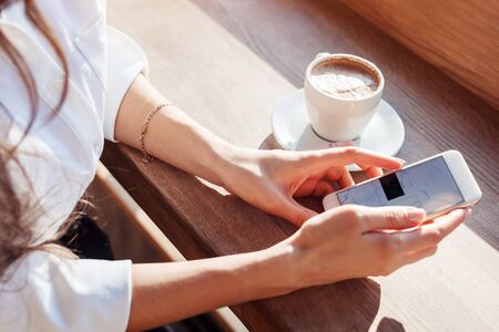 a girl in a cafe drinks coffee and uses the Internet in smart-phone. Workplace in a cafe. Sunlight comes from the window, toned photoの写真素材