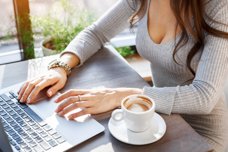 A sexy girl in a gray dress is working behind a laptop in a cafe, drinking coffee. The girl is typing on the laptop keyboard. Organization of the workplaceの写真素材