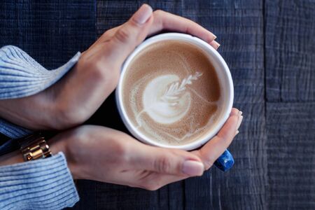 girl holds a blue cup of cappuccino coffee with latte art in hands on a wooden tableの写真素材