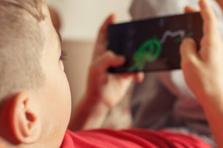 A boy lies, holds a mobile phone in his hands and plays a gameの写真素材