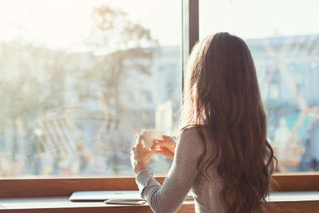 the woman in the cafe sits at a table, drinks coffee and looks at the panorama of the city outside the window. A girl in a cafe on a sunny morningの写真素材