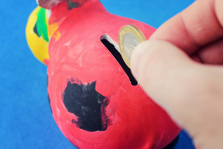 The hand drops the coin into the coin box. The concept of saving money. red piggy bank, painted by a child on a blue backgroundの写真素材