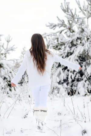 A girl with long hair in a white sweater is happy in a snowy forest. A girl is walking with her back hugging the wood with her handsの写真素材