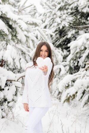 attractive girl in a white warm sweater in a winter forest holds a heart from the snowの写真素材