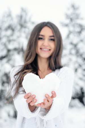 a young woman in a blue dress is holding a heart from the snow in a winter fir forest. Focus on the heart. The girl gives her heartの写真素材