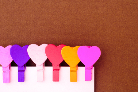 Small colored hearts hold a paper note on a brown background. The concept of a note to the day of St. Valentineの写真素材