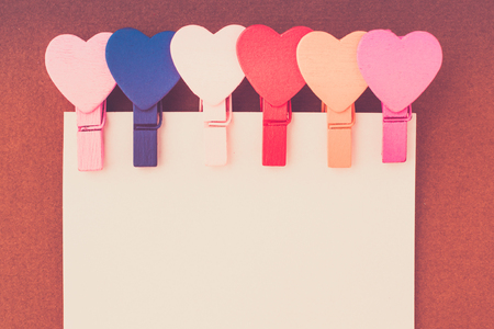 Small colored hearts hold a paper note on a brown background. The concept of a note to the day of St. Valentineの写真素材