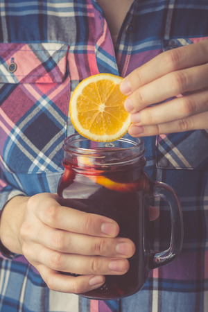 A young woman in a shirt holds a mug of homemade mulled wine in her hands, dips a slice of lemon into a cup. Toned photoの写真素材