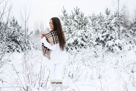 Attractive young woman in a white jumper and jeans wrapped in a scarf walking through the winter forest and enjoying lifeの写真素材