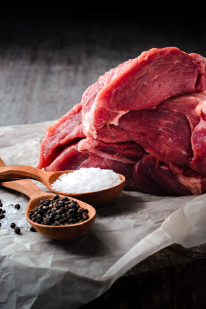 A large piece of pork meat, salt and pepper in wooden spoons on a wooden table on a dark background. Vertical compositionの写真素材