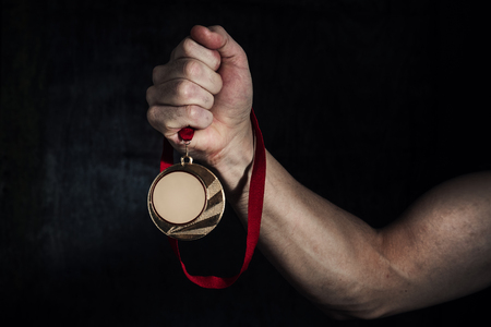 A dirty man's hand holds a gold medal on a dark background. Award of victory. The concept of successの写真素材