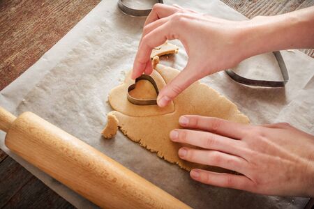 woman with her hands carves ginger cookies in the form of hearts with a metal shape on a wooden tableの写真素材