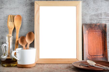 An empty wooden frame with isolated white copy space and kitchen accessories on the table. Mock up for designの写真素材