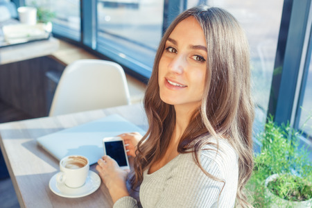 Attractive young woman uses a laptop and mobile phone in a cafe. Beautiful girl looks at the cameraの写真素材