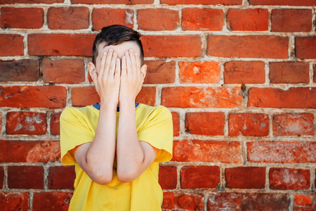 Boy in a yellow T-shirt covers his face with his hands against a brick wallの写真素材
