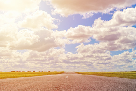 asphalt road through the green field and clouds on blue sky in summer day, toned photoの写真素材