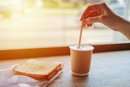 Breakfast in cafe with cup of coffee and toast. Woman's hand stirs coffee in paper cupの写真素材