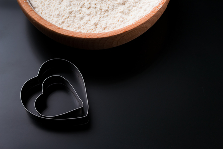 Dough cutter in shape of heart and bowl with flour on black table. Valentines Day baking backgroundの写真素材